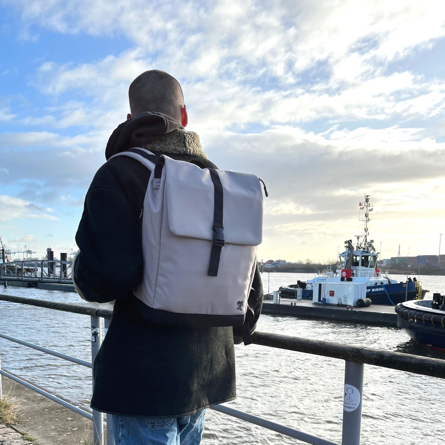 Junge Mann steht an der Elbe und traegt den FUCHS und REBELL MATS Daypack in beige