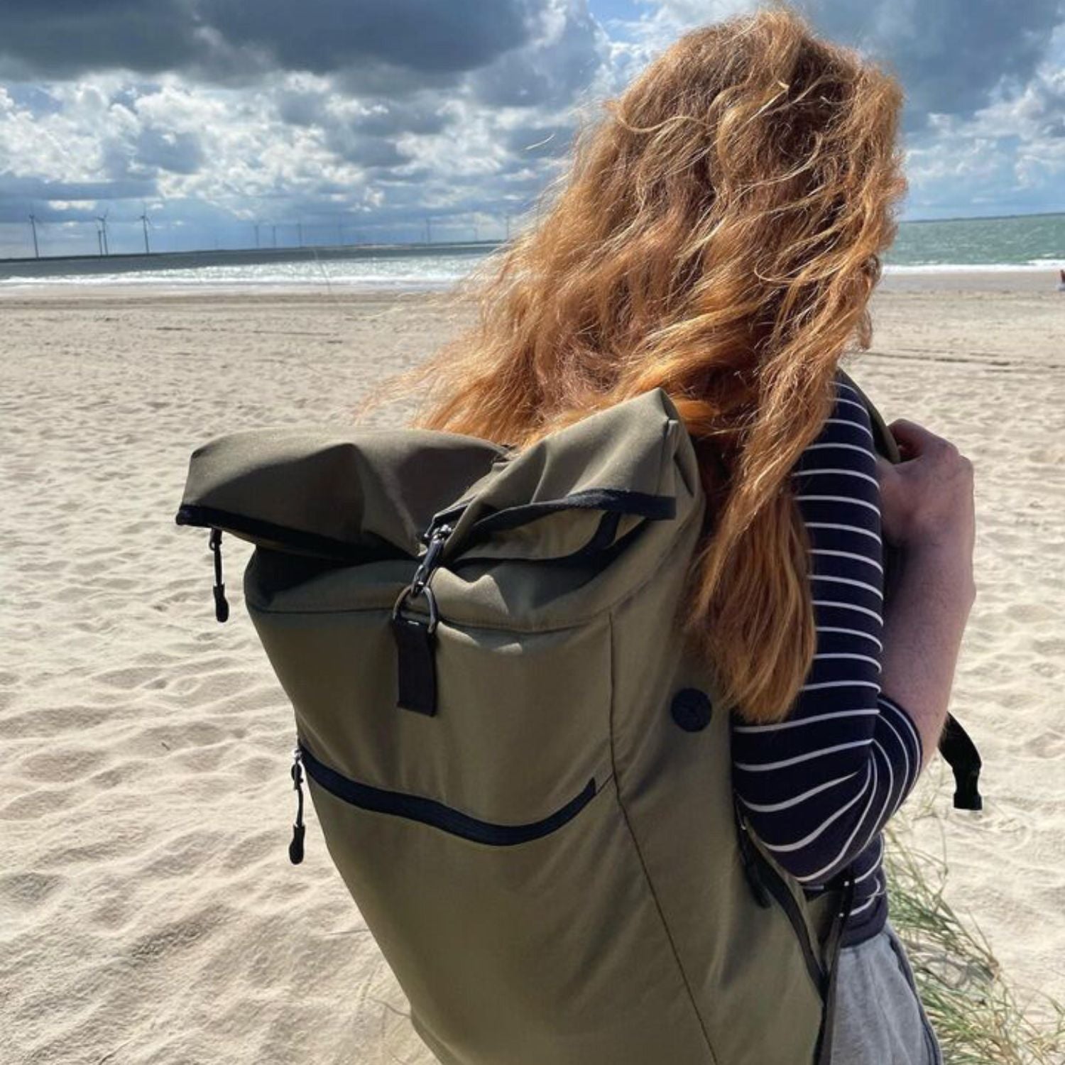 Junge Frau geht am Strand spazieren und traegt den FUCHS und REBELL PIET Rolltop Rucksack in gruen.
