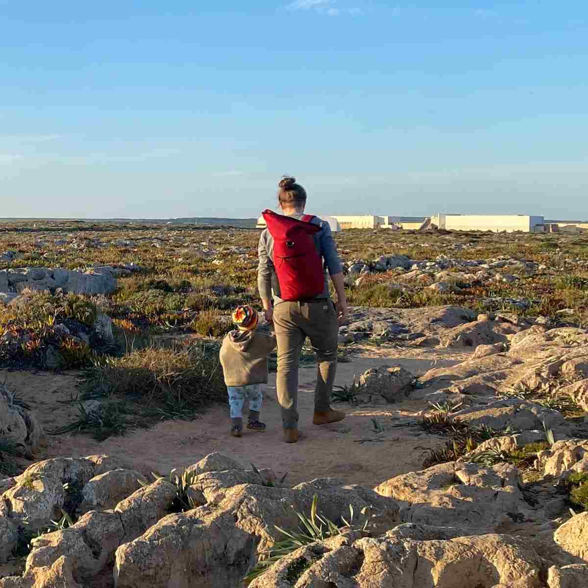 Mann geht mit seinem Kind am Strand spazieren und traegt den roten FUCHS UND REBELL PIET Rolltop Rucksack