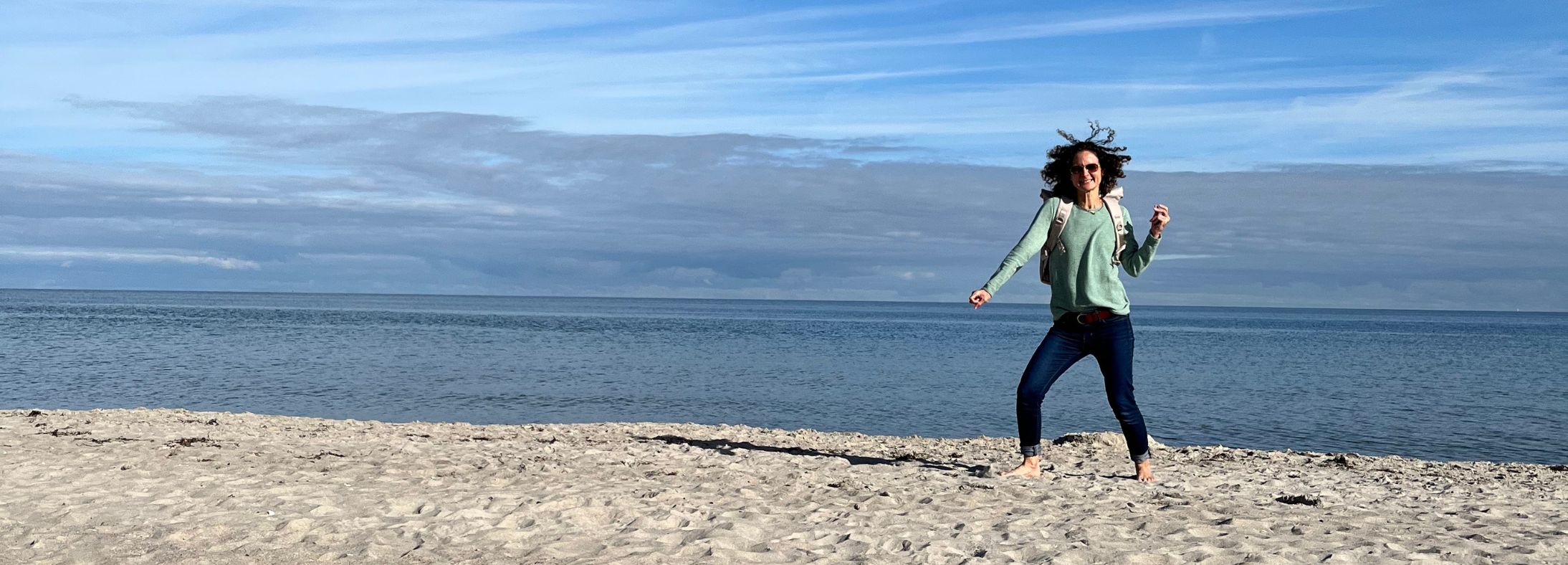 Eine Junge Frau springt am Strand in die Höhe und traegt den beigefarbenen FUCHS und REBELL PIET Rolltop Rucksack