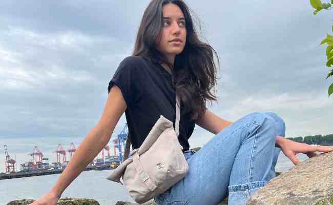 Frau sitzt auf Steinen am Strand und traegt die LUCA Crossbody Tasche in beige