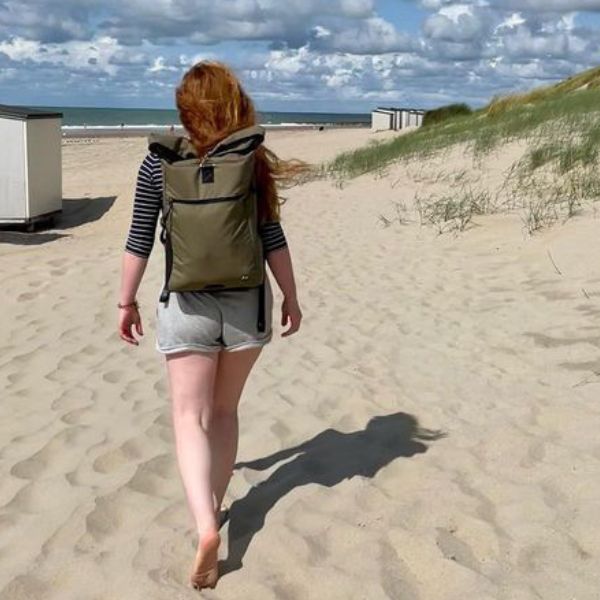 Junge Frau geht am Strand spazieren und traegt den gruenen FUCHS und REBELL PIET Rolltop Rucksack
