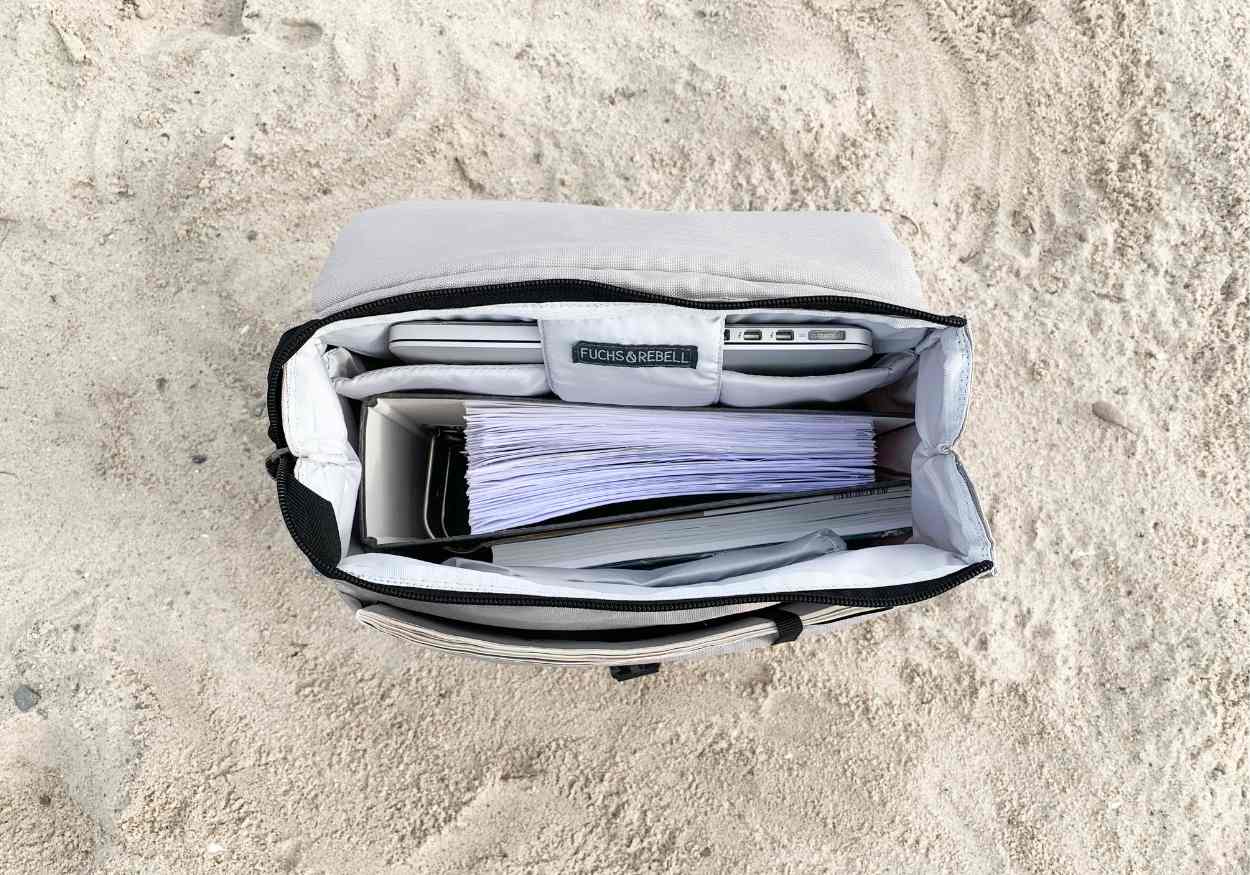 Innenansicht von dem vollgepackten FUCHS und REBELL Rucksack MATS in beige am Strand. Das Foto zeigt, dass sowohl ein DIN A4 Ordner als auch ein Laptop und viele andere Wertsachen Platz haben.