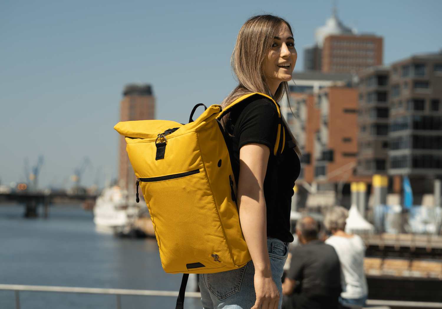 Eine junge Frau geht im Hamburger Hafen spazieren und traegt den senfgelben FUCHS und REBELL Rolltop Rucksack PIET.