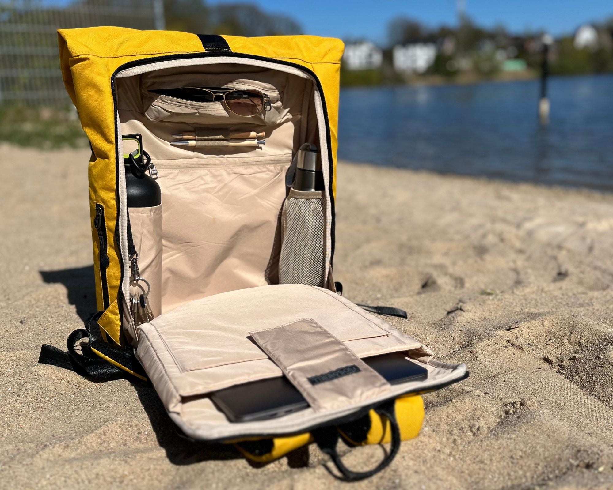 Der senfgelbe FUCHS UND REBELL PIET ROLLTOP RUCKSACK steht komplett offen am Strand und zeigt das Innenleben mit den vielen Taschen.
