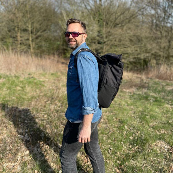Gut aussehender Mann trägt bei strahlendem Sonnenschein den schwarzen FUCHS und REBELL PIET Rolltop Rucksack und geht im Wald spazieren.