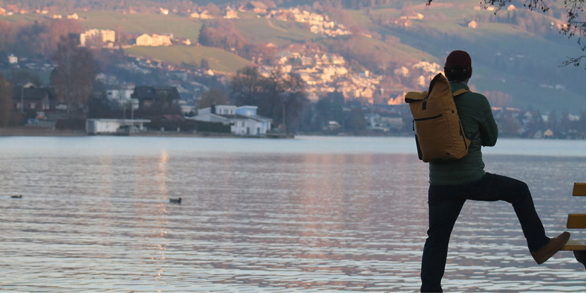 Ein Mann steht bei Sonnenuntergang  am Zuger See und traegt den senfgelben FUCHS und REBELL Rucksack