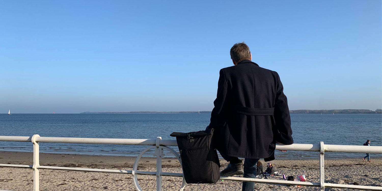 Ein Mann sitzt am Travemuender Strand, schaut auf das Meer und haelt den schwarzen FUCHS und REBELL PIET Rolltop Rucksack in der Hand