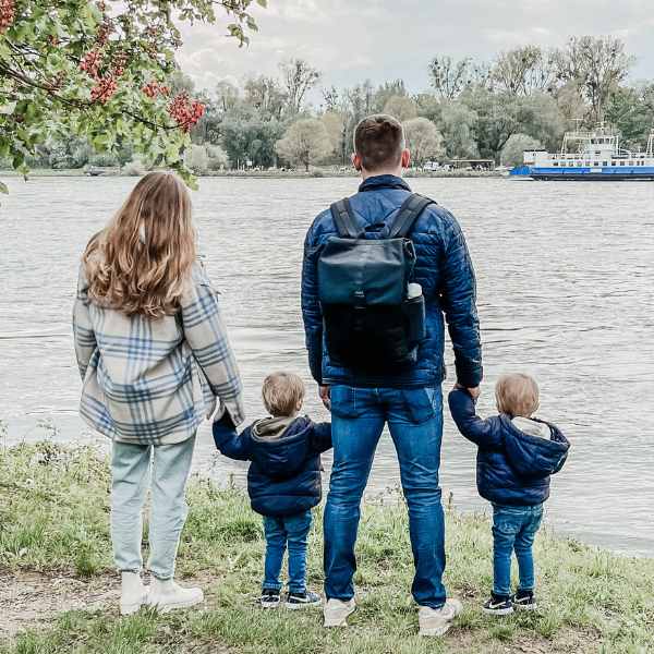 Junge Familie steht mit ihren Zwillingen am See. Der Vater traegt den schwarzen FUCHS & REBELL MATS Daypack.