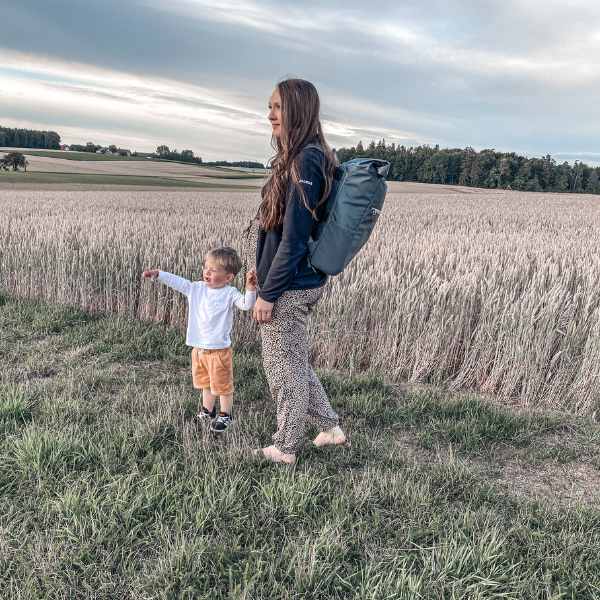 Junge Mutter steht mit ihrem Kleinkind am Weizenfeld und traegt den dunkelgrauen FUCHS & REBELL PIET Rolltop Rucksack