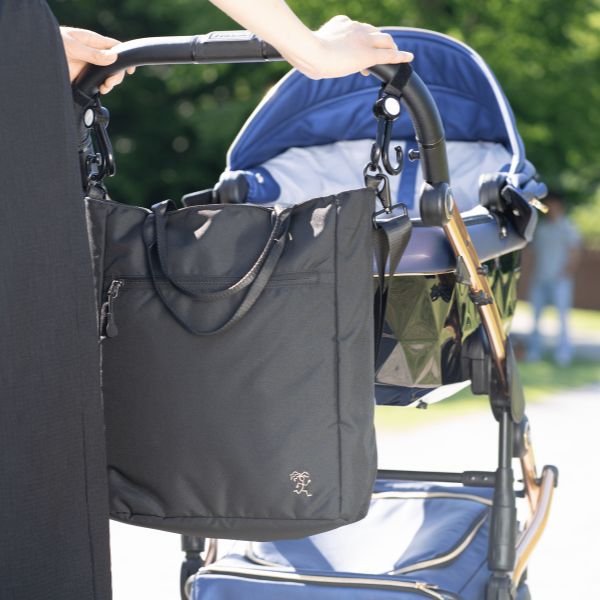 unge Mutter geht mit ihrem Kind im Kinderwagen spazieren. Die FUCHS & REBELL JONA Umhängetasche hängt als Wickeltasche an einem Kinderwagen.