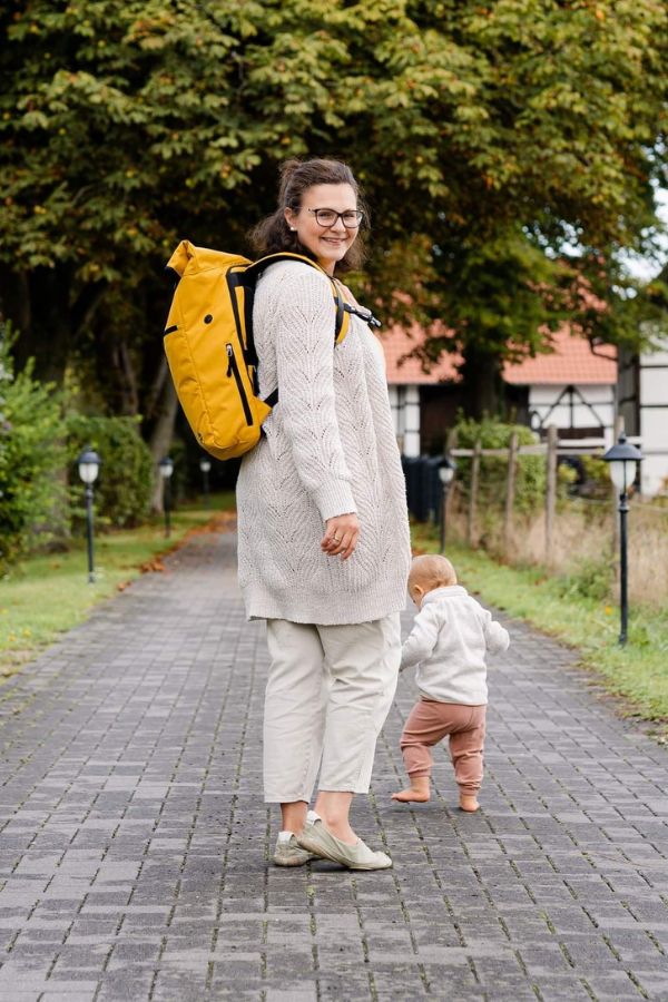 Junge Mutter lächelt und traegt ihren senfgelben FUCHS & REBELL PIET Rolltop Rucksack und geht mit ihrem Saeugling spazieren.