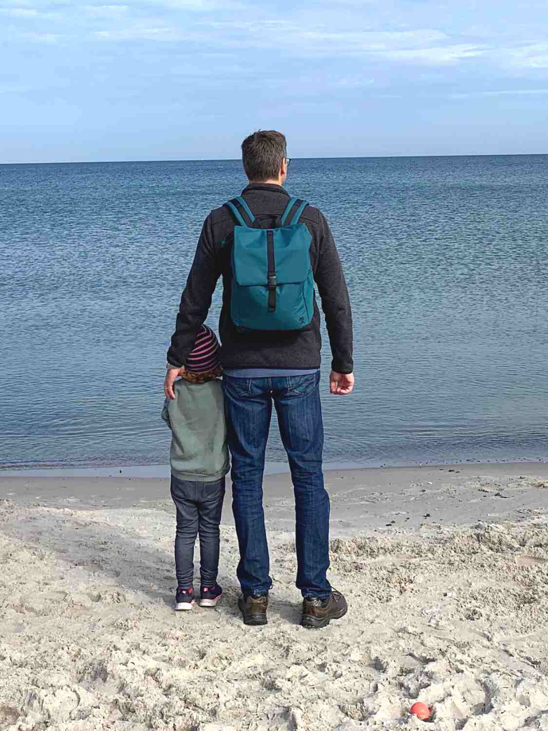 Junger Mann steht mit seiner Tochter am Meer, traegt den petrolblauen FUCHS und REBELL MATS Daypack Rucksack und schaut auf das Meer