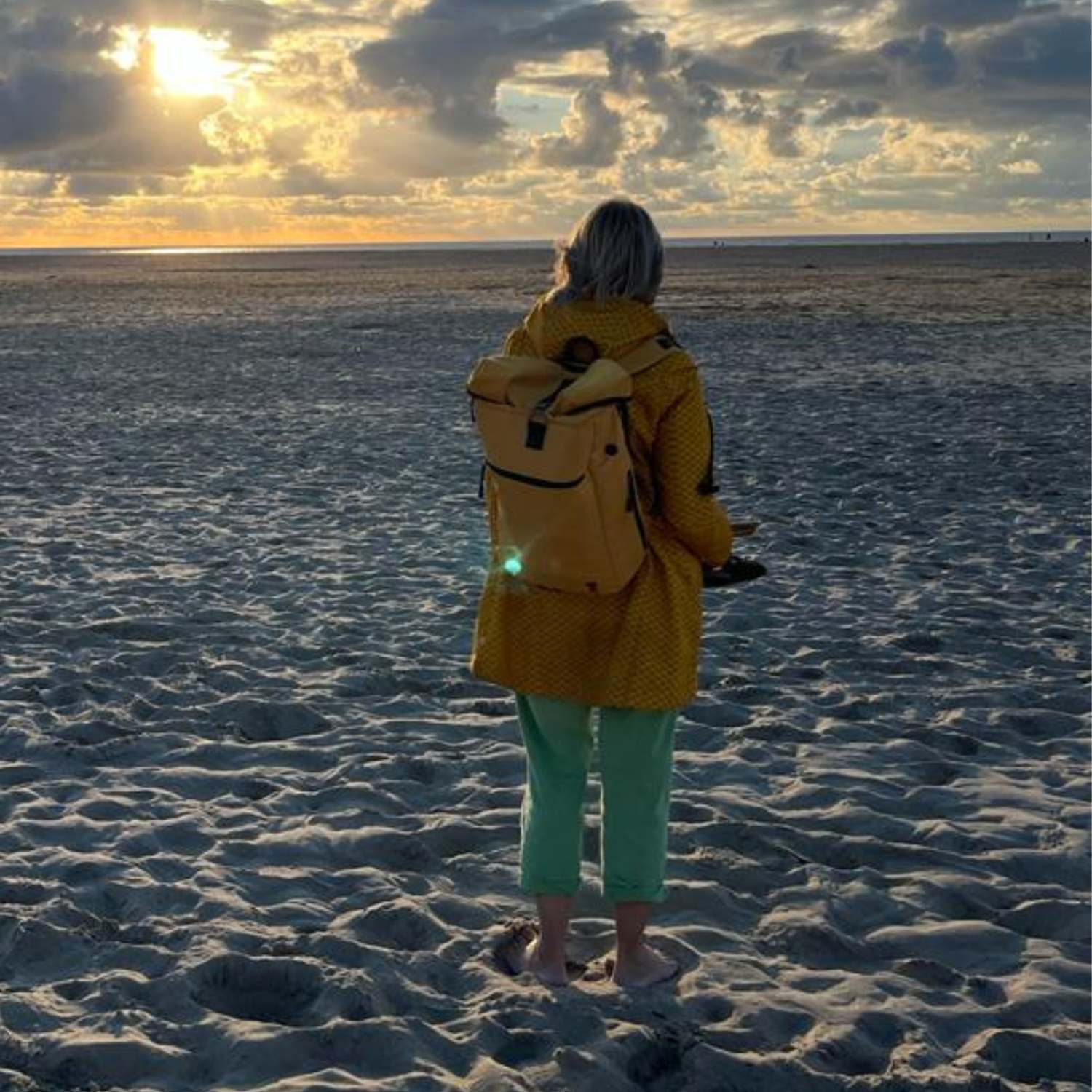 Junge Frau geht am Strand spazieren und traegt den FUCHs und REBELL PIET Rolltop Rucksack in senfgelb