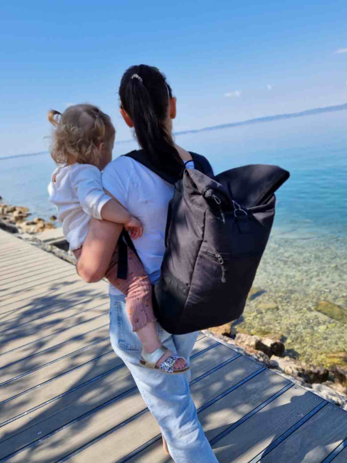 Junge Frau traegt ihr Baby und den FUCHS und REBELL schwarzen PIET Rolltop Rucksack am  Strand spazieren