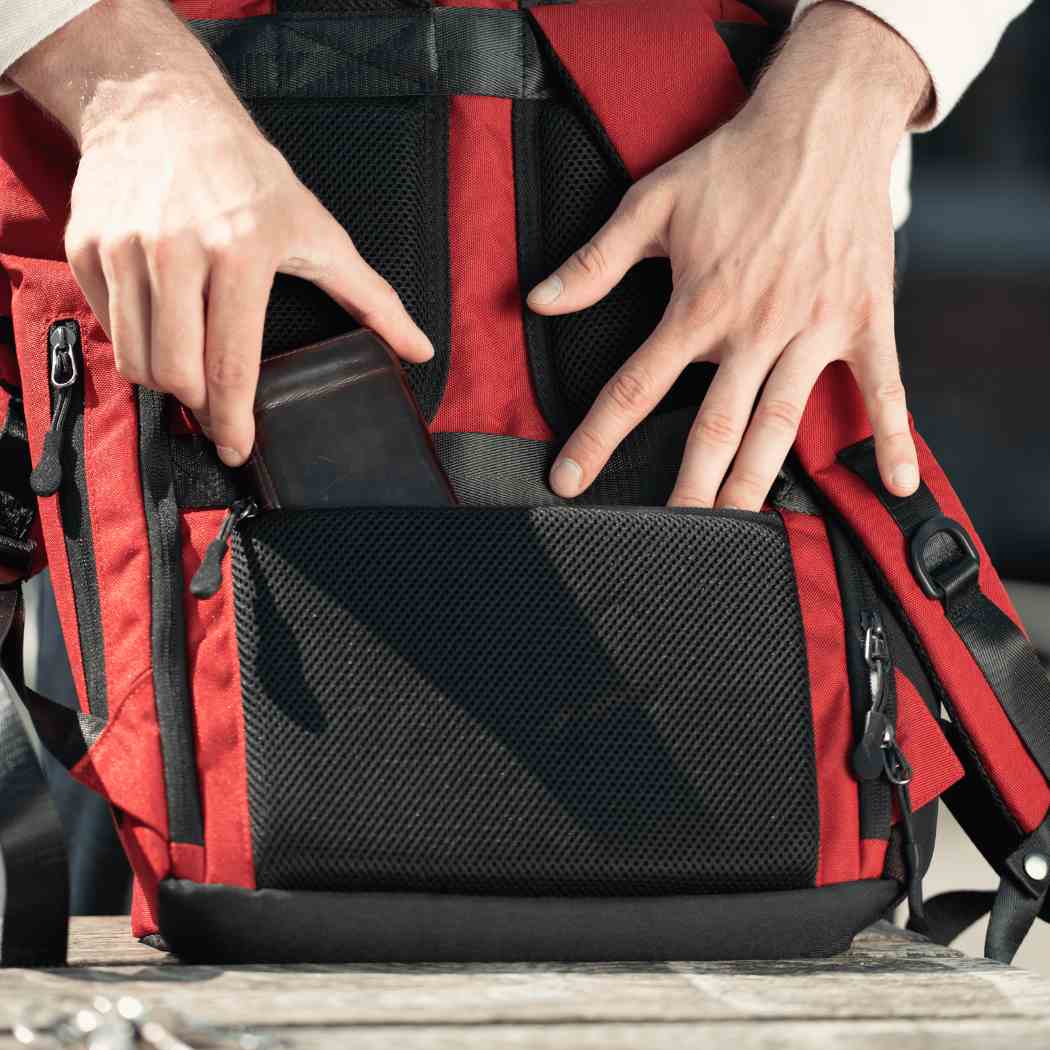 Mann zeigt das Anti-Diebstahl Fach auf der Rueckseite des roten FUCHS UND REBELL PIET Rolltop Rucksack 