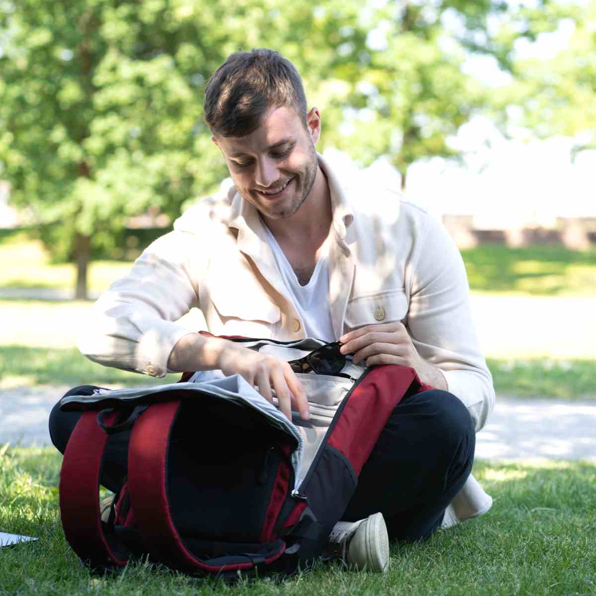 Mann hat den FUCHS UND REBELL PIET Rolltop Rucksack in rot auf dem Boden durch den hinteren Reissverschluss geöffnet und holt seine Brille raus.