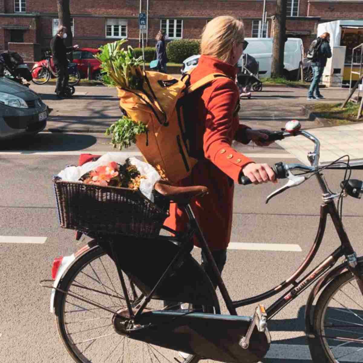 Frau schiebt ihr Fahrrad und traegt ihre Einkaeufe in ihrem senfgelben FUCHS UND REBELL PIET Rolltop Rucksack