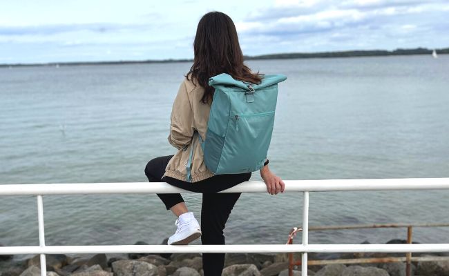 Junge Frau sitzt auf einem Geländer und schaut auf die Ostsee wärend sie den FUCHS und REBELL PIET Rolltop Rucksack in mint blau traegt.