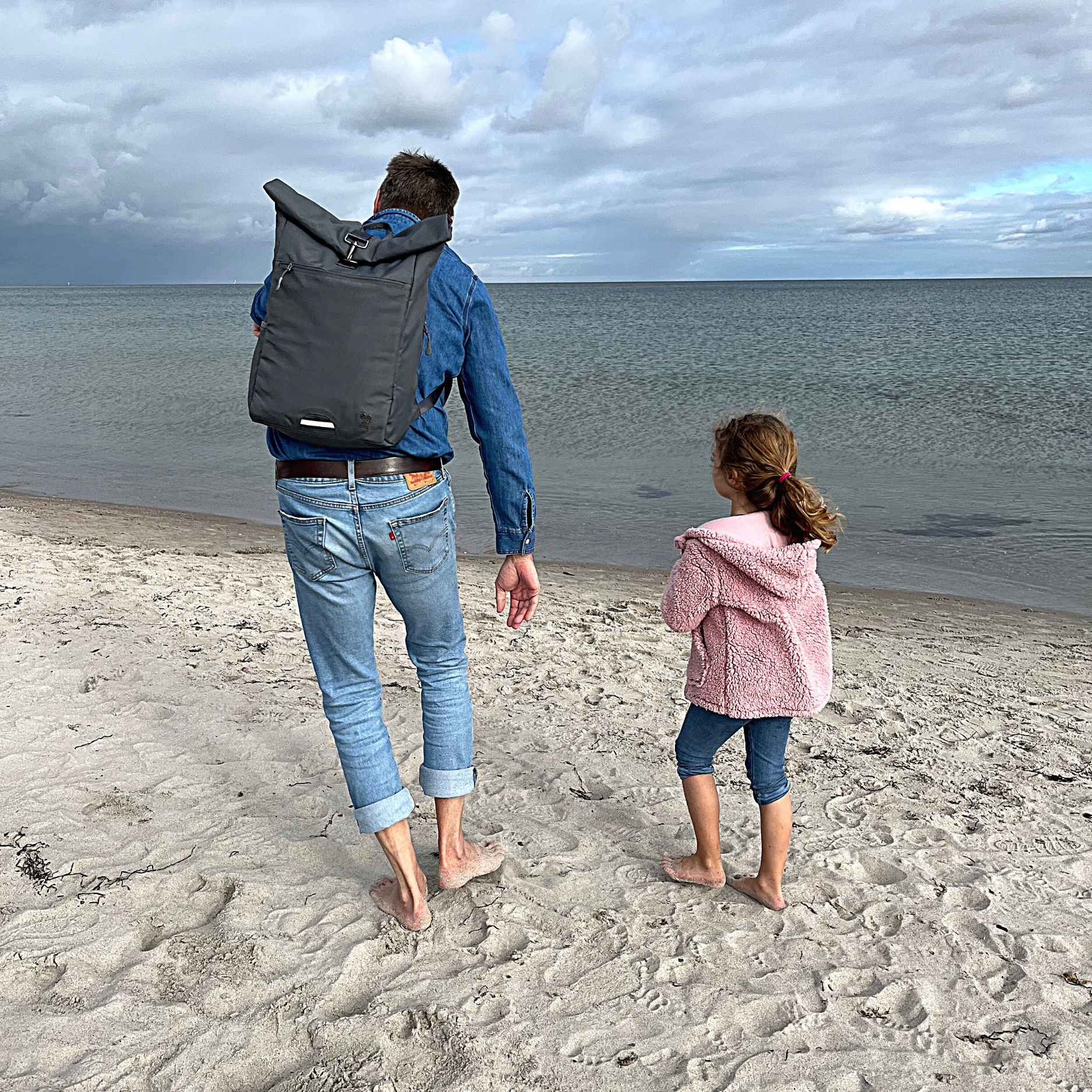 Mann traegt den FUCHS und REBELL PIET Rolltop Rucksack in dunkelgrau mit Kind am Strand