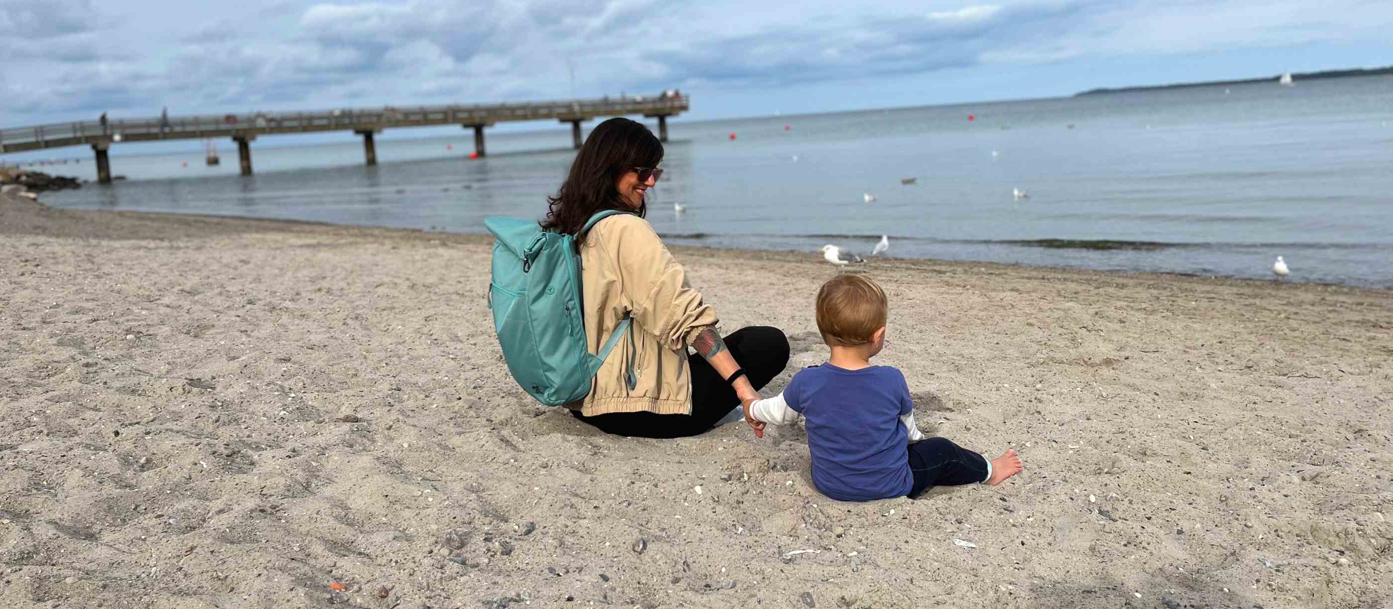 Eine Mutter sitzt mit ihrem Baby am Strand und traegt ihren FUCHS und REBELL PIET Rolltop Rucksack in mintblau auf dem Ruecken. 