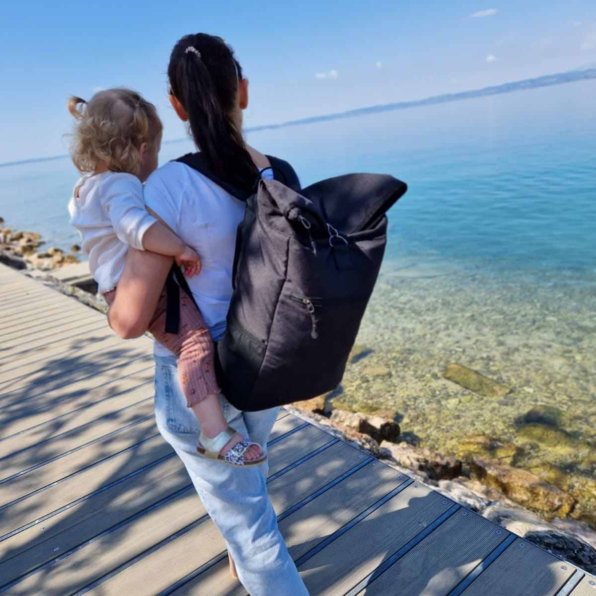 Eine Frau traegt ihr Baby auf dem Arm, geht auf einem Steg am Meer spazieren und traegt den schwarzen FUCHS und REBELL PIET Rolltop Rucksack.