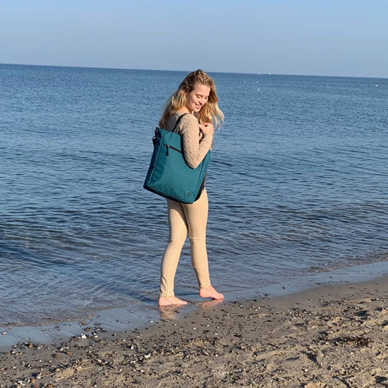 Eine junge Frau geht am Strand spazieren und zeigt dass man die petrolblaue JONA Umhaengetasche von FUCHS und REBELL über der Schulter tragen kann. 