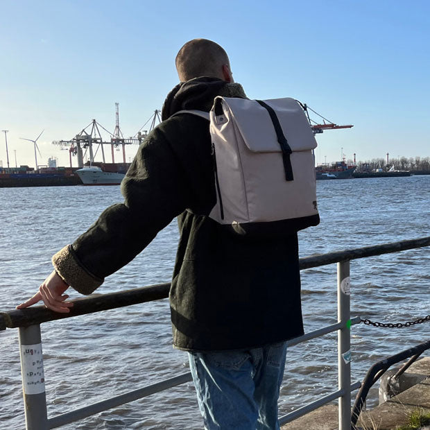 Junger Mann steht an der Elbe und trägt den beigefarbenen FUCHS und REBELL MATS Rucksack