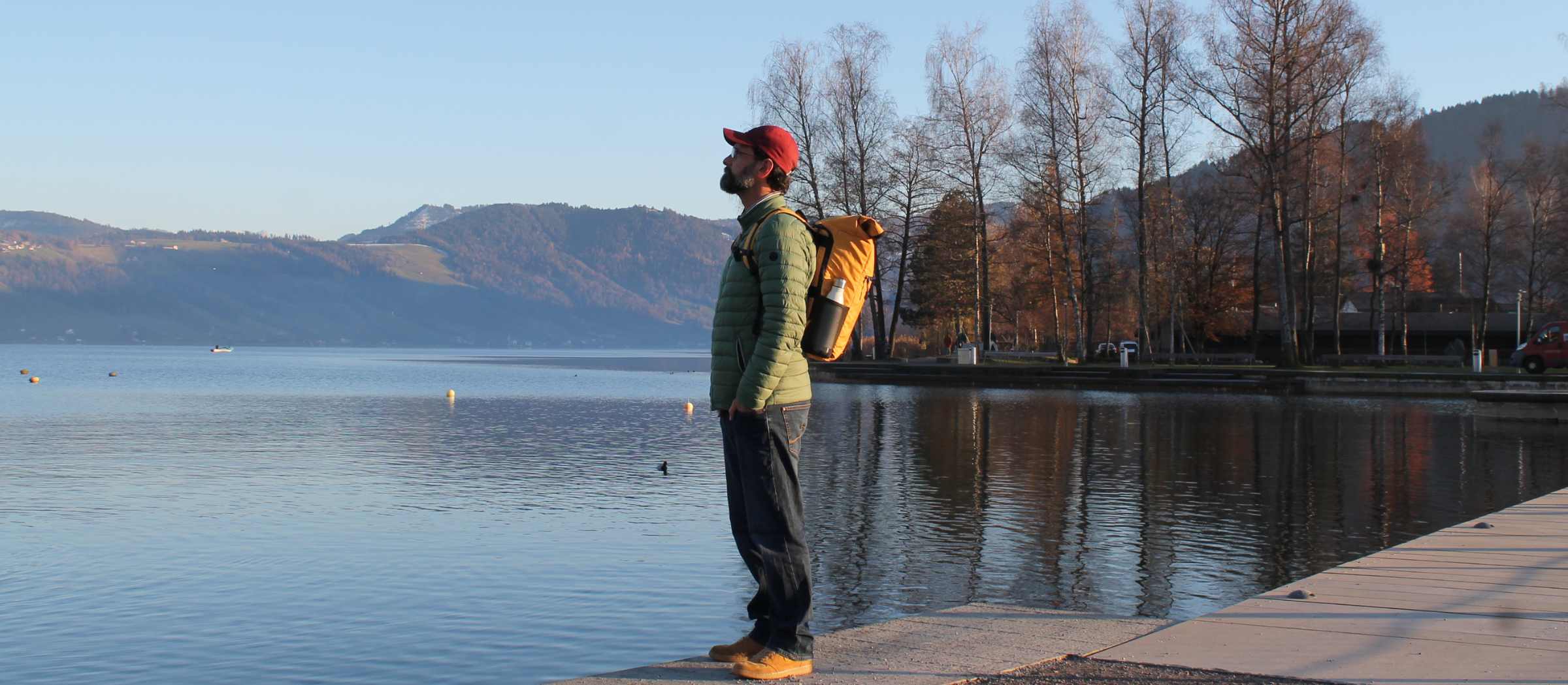 Junger Mann steht am See, traegt den gelben FUCHS und REBELL PIET Rolltop Rucksack und schaut auf den See
