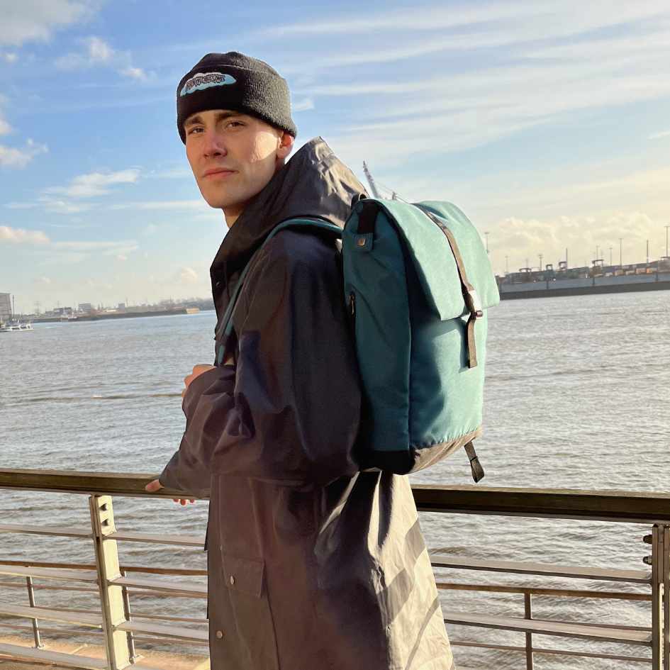 Junger Mann steht an der Elbe und traegt den petrolblauen FUCHS und REBELL MATS Rucksack