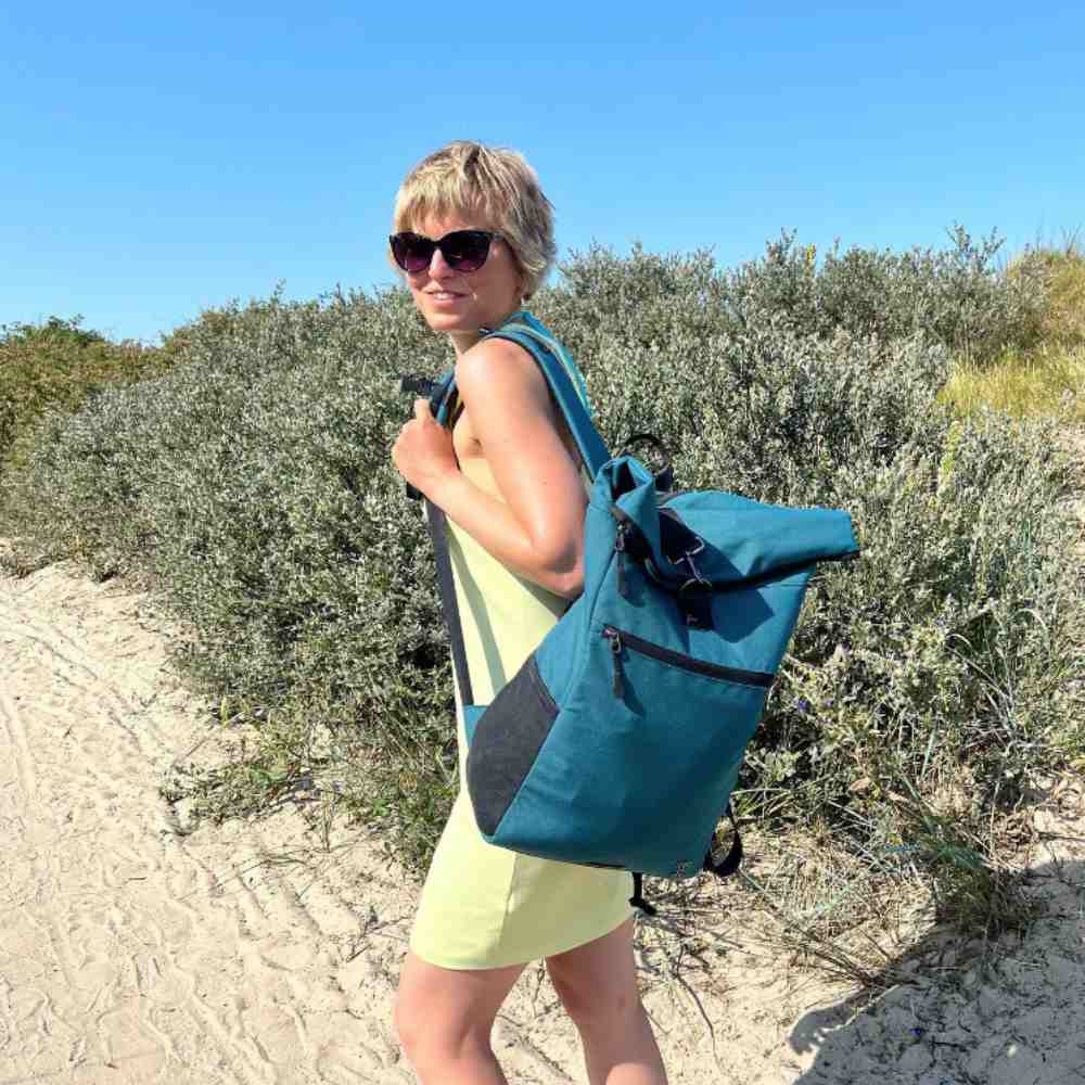 Gutaussehende Frau geht mit dem petrolblauen PIET Rolltop Rucksack am Strand spazieren.