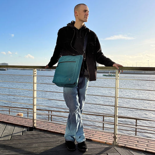 Junger Mann steht an der Elbe und trägt die petrolblaue FUCHS und REBELL JONA Umhaengetasche als Cross-Body-Tasche
