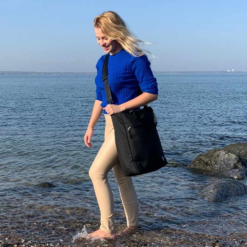 Junge Frau geht am Strand spazieren und traegt die schwarze FUCHS und REBELL JONA Umhaengetasche.