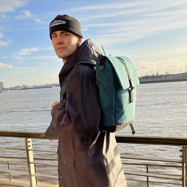 Junger Mann steht an der Elbe und trägt den petrolblauen FUCHS und REBELL MATS Rucksack