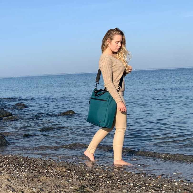 Junge Frau geht am Strand spazieren und traegt die petrolblaue FUCHS und REBELL JONA Umhaengetasche.
