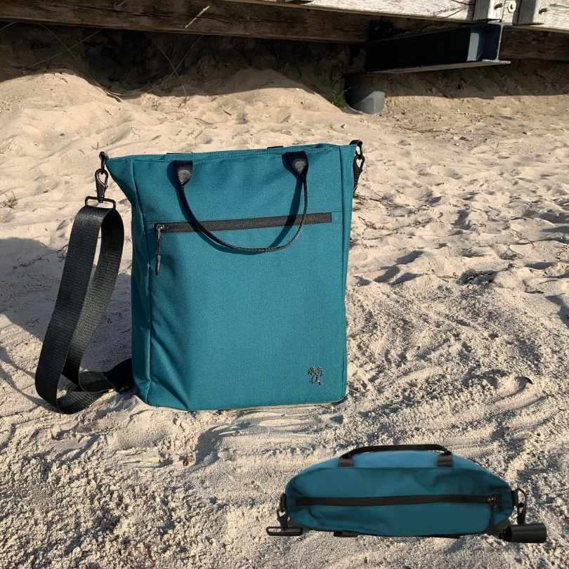 Die nachhaltige FUCHS und REBELL Umhaengetasche JONA in petrolblau steht am Strand. Zusätzlich zeigt die ANsicht von oben dass alle Außentaschen mit hochwertigen Reissverschlüssen versehen sind.