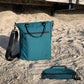 Die nachhaltige FUCHS und REBELL Umhaengetasche JONA in petrolblau steht am Strand. Zusätzlich zeigt die ANsicht von oben dass alle Außentaschen mit hochwertigen Reissverschlüssen versehen sind.